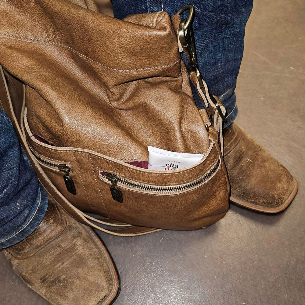 Leather crossbody bag in brown with an open zipper, revealing interior items, paired with cowboy boots.