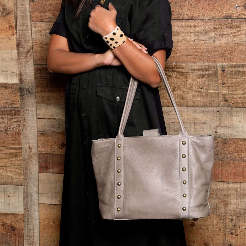Person holding a gray leather tote bag against a wooden wall