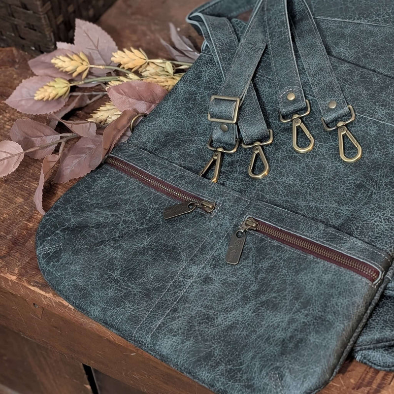 A dark blue/grey textured full-grain leather bag with antique brass hardware, shown resting on a rustic wooden surface next to dried autumn leaves.
